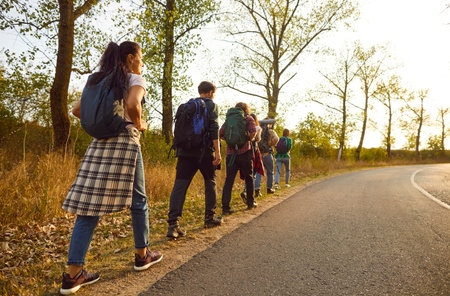Group of tourists hiking or trekking on warm September day. View from behind team of young people with backpacks walking together on roadside along highway lined with green treesの写真素材