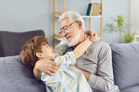 Portrait of cheerful senior grandfather with happy boy grandson sitting on sofa at home hugging and smiling. Elderly man having fun with child indoors enjoying time together. Family and love concept.の写真素材