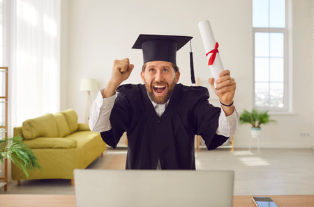 Happy excited student man wearing graduate gown and hat sitting at desk with diploma in hands in front of laptop making yes gesture during video call. University education and online degree conceptの写真素材