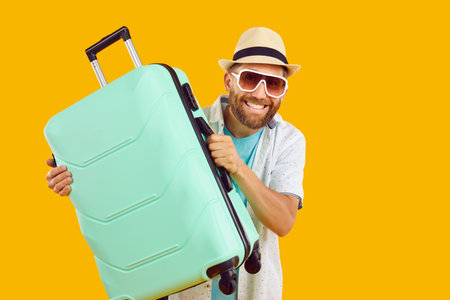Happy male tourist with packed suitcase ready for summer holiday. Joyful bearded man in summer hat and sunglasses holding green bag isolated on yellow orange color background. Travel, vacation conceptの写真素材