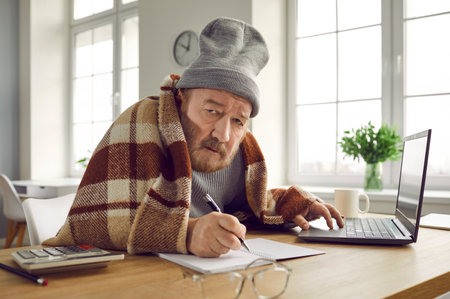 Portrait of a senior man, working in the cold at home, sitting at the table wrapped in a blanket and a hat, typing on his laptop computer and writing in a notebook. Heating problems.の写真素材