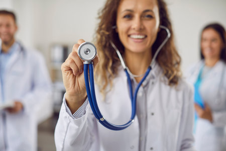 Close up portrait of a young happy smiling pretty female doctor in white medical uniform standing with stethoscope in clinic. Confident woman physician looking cheerful at camera. Selective focus.の写真素材