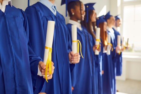 Group of happy adult diverse multiethnic college or university students in traditional blue hats and robes attending indoor graduation ceremony, standing in row, and holding diploma scrolls in handsの写真素材