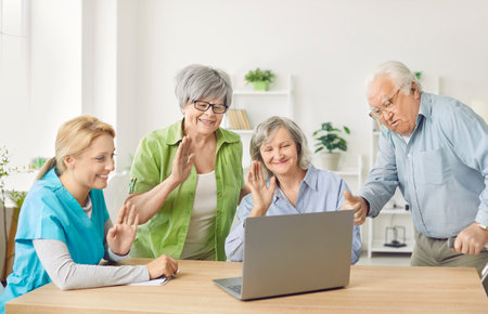 Group of happy senior people sitting at table in nursing home with a young friendly nurse, waving hand having online video call using laptop. Retirement and communication technology concept.の写真素材