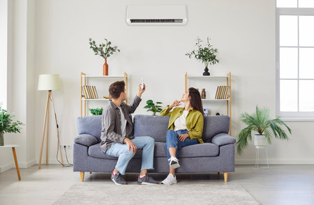 Young married couple relaxing sitting on couch and enjoying their new modern air conditioning system at home. The man using the remote AC control to turn on air conditioner or regulate temperature.