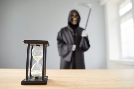 Hourglass standing at desk indoors with a man wearing Halloween grim reaper death costume holding...