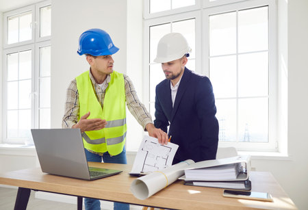 Two men in construction helmets discuss the master plan in a room, close-up. Concept design...