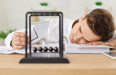 Tired businessman sitting at work desk with Newtons cradle pendulum at foreground, putting head on...
