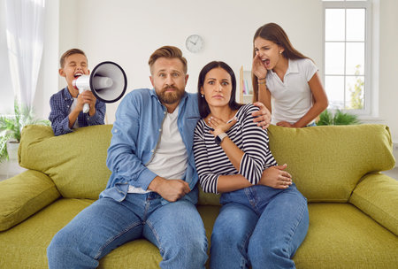 Tired exhausted family mother and father sitting on couch at home while noisy kids daughter and son shouting in loudspeaker in background. Problems of parenthood and need repose concept.の写真素材