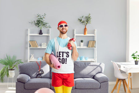 Portrait of funny young man wearing sportswear holding yoga mat and dumbbells in hands and looking...