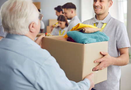 A smiling volunteer guy gives an elderly man a box of things. Concept for caring for the elderly and displaced people. Premises of a charitable foundation for supporting migrants and refugeesの写真素材