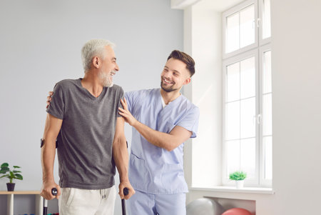 Happy health care worker with elderly man smiling and hugging in rehabilitation clinic. Young cheerful nurse helping senior patient to walk with his crutches. Physiotherapist helping old man in rehabの写真素材
