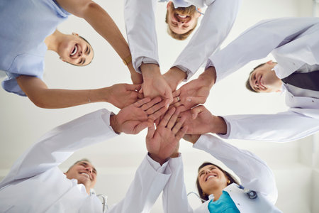 Bottom view of doctors in white uniform standing in a circle in hospital or clinic putting their arms in a pile. Stack of medical staff hands. Concept of unity, partnership, teamwork and health careの写真素材