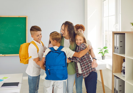 Portrait of a happy schoolchildren group with backpacks standing with their female friendly teacher meeting after holidays in classroom at school and hugging. Back to school concept.の写真素材