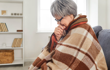Sad sick gray-haired senior woman sitting on sofa at home wrapped in a blanket feeling unhealthy with influenza. Elderly retired person freezing in winter in chilly room with low temperature.の写真素材