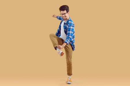 Happy young man is dancing, having fun and fooling around. Bearded 20-year-old man with glasses, dressed in plaid blue shirt and beige trousers. Full-size photo on an isolated beige background.の写真素材