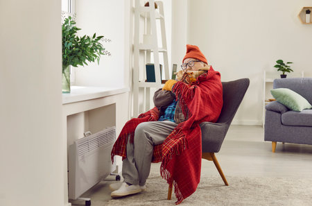 Frozen senior man battles the winter chill in living room at home, clad in outerwear and swathed in a blanket. Despite the presence of a heater, the room carries a hint of frosty discomfort.の写真素材