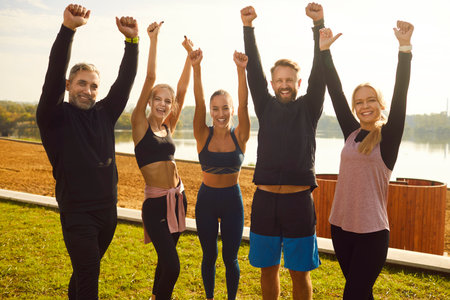 Portrait of a happy satisfied sporty people in sportswear standing with hands up in fist like a winners on lake shore after sport exercises in park. Outdoors training and fitness in nature concept.の写真素材