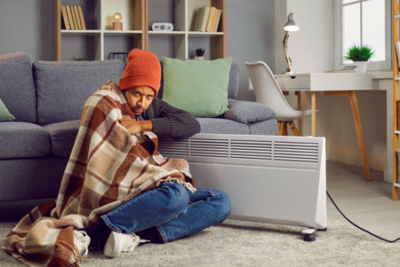 Disappointed man covered with blanket sitting on floor beside electric heater. Upset warmly dressed...
