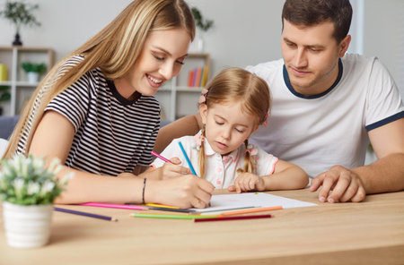 Portrait of happy family sitting at desk and drawing together. Smiling mother, father and their daughter painting picture with colorful pencils, having fun, developing creativity of preschool child.の写真素材