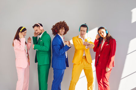 Creative stylish people in headphones seem to be moving to beat of their favorite tunes. Group of young energetic people in colorful suits stand in row and dance immersed in their own world of music.の写真素材