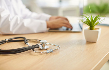 Close up of male doctor sitting at the desk on workplace in medical clinic with stethoscope in foreground. Man physician in white uniform working using laptop. Health care and medicine concept.の写真素材