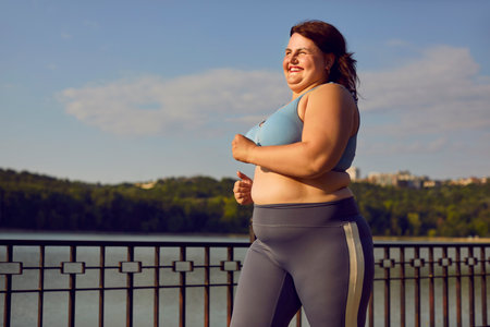 Happy cheerful joyful smiling fat overweight chubby big large plump stout young woman in a sports bra and yoga pants running on a city river bridge during her outdoor jogging workout in summerの写真素材
