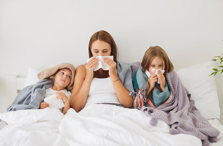 Portrait of young sick family of mother and two little children lying in bed covered in blankets with seasonal flu or cold. Woman with kids suffering from runny nose together at home.の写真素材
