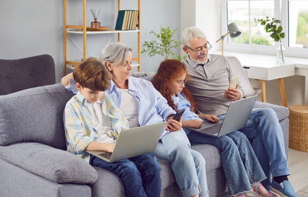 Happy grandparents with grandchildren girl and boy sitting on sofa using modern laptop and...
