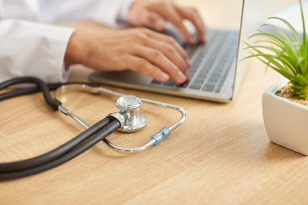 Close up shot of a stethoscope lying on a desk, with a doctor or nurse working online on a laptop in the background. Essential medical equipment used in hospital and clinic office.の写真素材