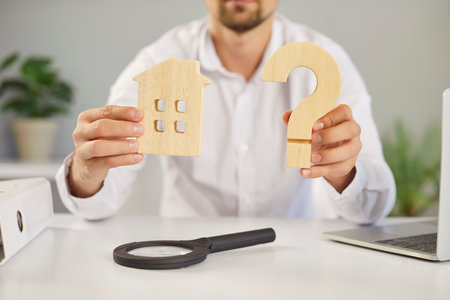 Estate agent, appraiser or mortgage broker sitting at an office table holding a wooden house symbol and question mark in his hands. Real estate, property, sale, help, advice conceptの写真素材