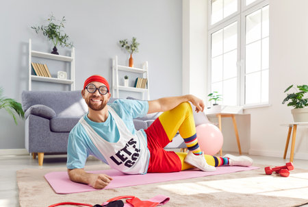 Portrait of funny young man wearing bright sportswear lying on floor on yoga mat and looking cheerful at camera in living room at home. Fitness, workout sport and home training concept.の写真素材