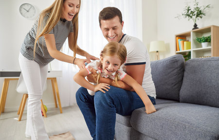 Happy parents playing together with young daughter while sitting on the couch at home. The family shares joyful leisure time, strengthening bond with affection, love, and childhood fun.の写真素材