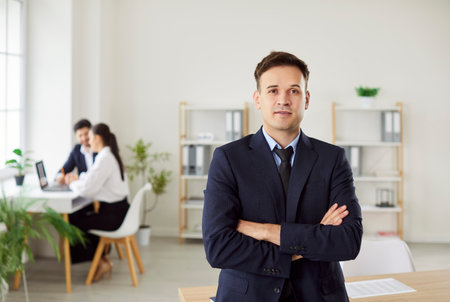 Portrait of confident businessman, company employee, CEO wearing black suit, standing with arms crossed and looking at camera, with team of colleagues working on background in modern office workplace.の写真素材