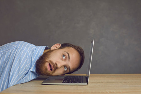 View from the side of a focused man who puts his ear to the laptop listening intently to its sounds. Bearded man in a shirt sits at a table and puts his head to the computer and looks up thoughtfully.の写真素材