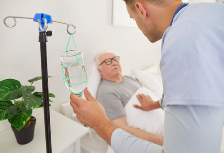 Caring young male nurse checking condition of senior patient during IV drip infusion in rehabilitation clinic. Friendly caregiver comforting elderly man lying in bed during vitamin therapy injection.の写真素材