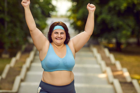 Happy young overweight woman having fitness workout in park. Cheerful beautiful fat chubby girl in sports bra and sweatband running down stone steps with her hands up, with green trees in backgroundの写真素材