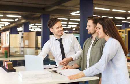 Young caucasian couple standing at building market or big shopping mall with male friendly salesman manager helping them to make purchase in hypermarket. Man seller consulting customersの写真素材