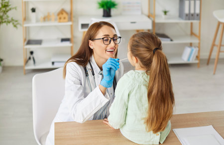 Female smiling pediatrician examining little girl patients throat in modern medical clinic on...