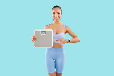 Sportswoman on blue background points finger at scales, close-up. Happy girl in sportswear controls...