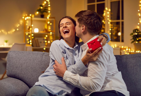 Happy husband surprises wife with a gift box at home living room, celebrating Valentine Day, anniversary or birthday. Love and joy shine during this special moment of holiday and affection.の写真素材
