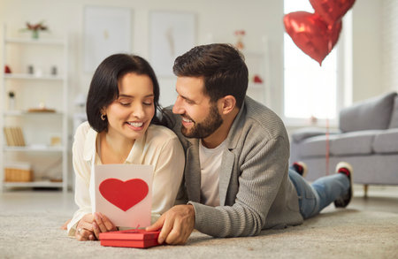 Happy couple unwraps a gift with a red heart on Valentine Day lying on floor. Celebrating love at home, they enjoy this romantic holiday together, surrounded by family and affection.の写真素材