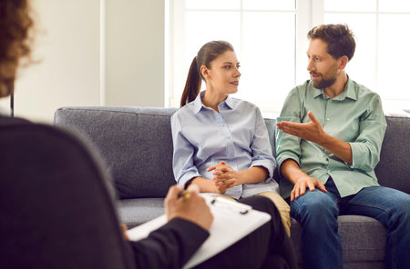 Consultation with family psychologist. Young man and wife are sitting on couch and talking to each other at psychotherapists appointment. Concept of marriage crisis, conflict and relationship problemの写真素材