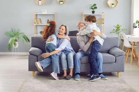 Grandparents and children having fun at weekend. Grandma and grandpa are happy to see kids. Grandmother and grandfather sitting on grey sofa and hugging grandchildren. People, family, love conceptsの写真素材