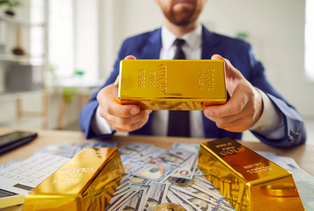 Rich man wearing business suit, holding a solid gold bar, surrounded by cash on table, symbolizing money wealth, finance, and business assets, while working in an office environment.の写真素材