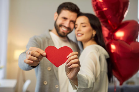 Young brunette man and woman holding heart in hands and hugging with red foil balloons in background celebrating holiday. Happy couple in love. Care, tenderness and Valentines day concept.の写真素材