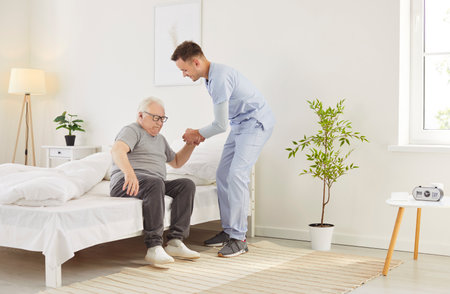 Caring male doctor or nurse helping a senior patient get out of bed after wake up in the morning at a nursing home. Compassionate healthcare, morning routines, and essential assistance.の写真素材