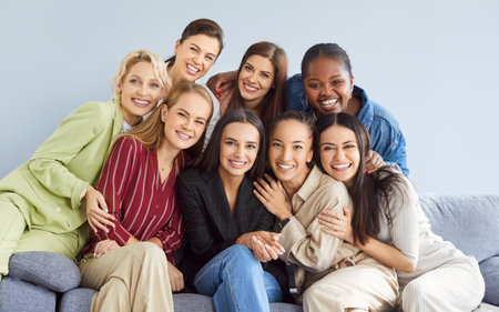 Portrait of group of female happy cheerful young diverse women friends coworkers or company employees hugging and looking at camera posing and smiling sitting on sofa on gray background.の写真素材