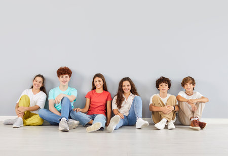 Group of happy students or friends sits together on the floor for a fun portrait on a grey background. The kids are enjoying time as a team, friendship and unity in this diverse and lively setting.の写真素材