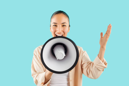 Studio shot of happy young Asian woman screaming in megaphone. Cheerful young girl isolated on bright blue background holding loud speaker in hand, looking at camera and talking with amplified voiceの写真素材